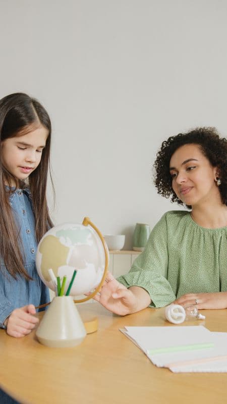 Fonoaudióloga e criança observando juntas um globo sobre a mesa, em atividade de aprendizado.