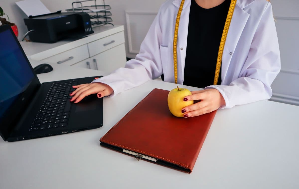 Nutricionista à mesa do consultório com laptop, agenda e uma maçã, organizando o trabalho.