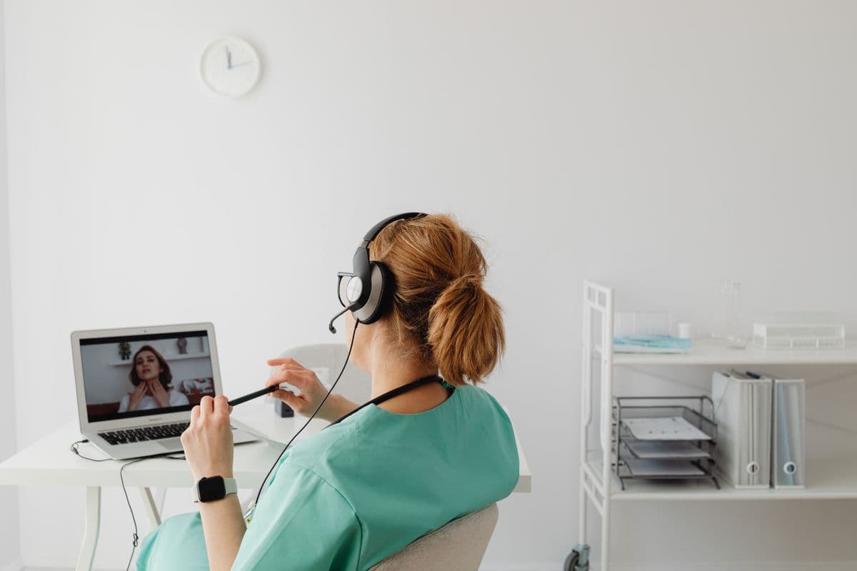 Profissional de saúde com headset atendendo uma paciente por vídeo no computador.